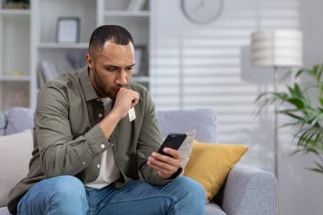 A concerned man in casual clothing sits on a couch, holding his phone and a credit card while looking intensely at the screen. The context is part of a website post entitled "When Home Turns Hostile".